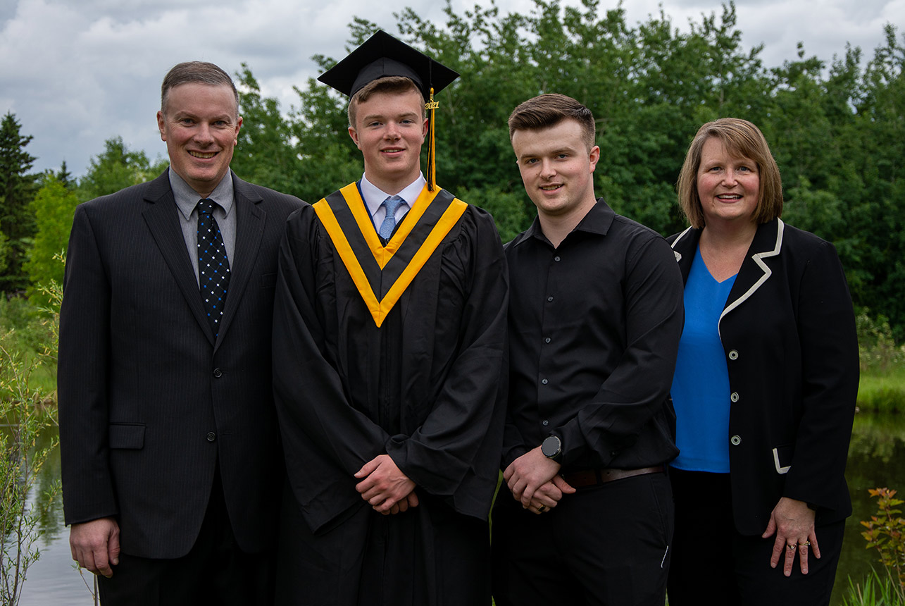 Graduating young man in cap and gown stands with family by pond surrounded by lush foliage.