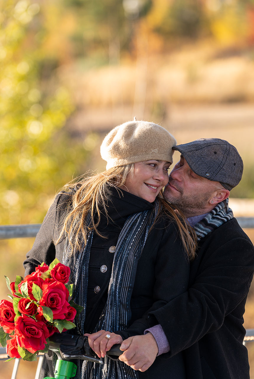 Woman in a wool beret smiles at her partner as he hugs her from behind, a bouquet of red roses resting on an electric scooter handlebar in warm autumn light