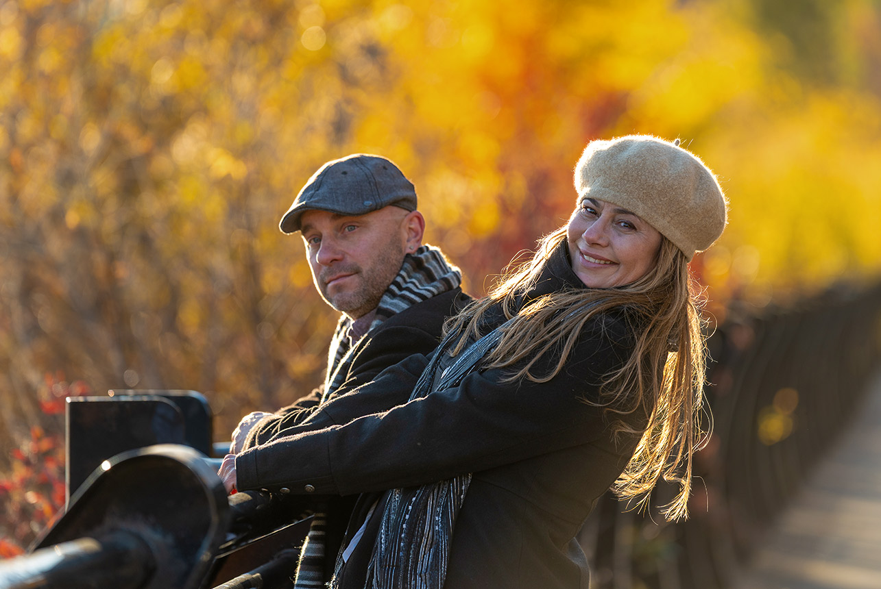 Couple lean on a riverside railing at Louise McKinney Riverfront Park, bathed in warm autumn light with golden trees behind