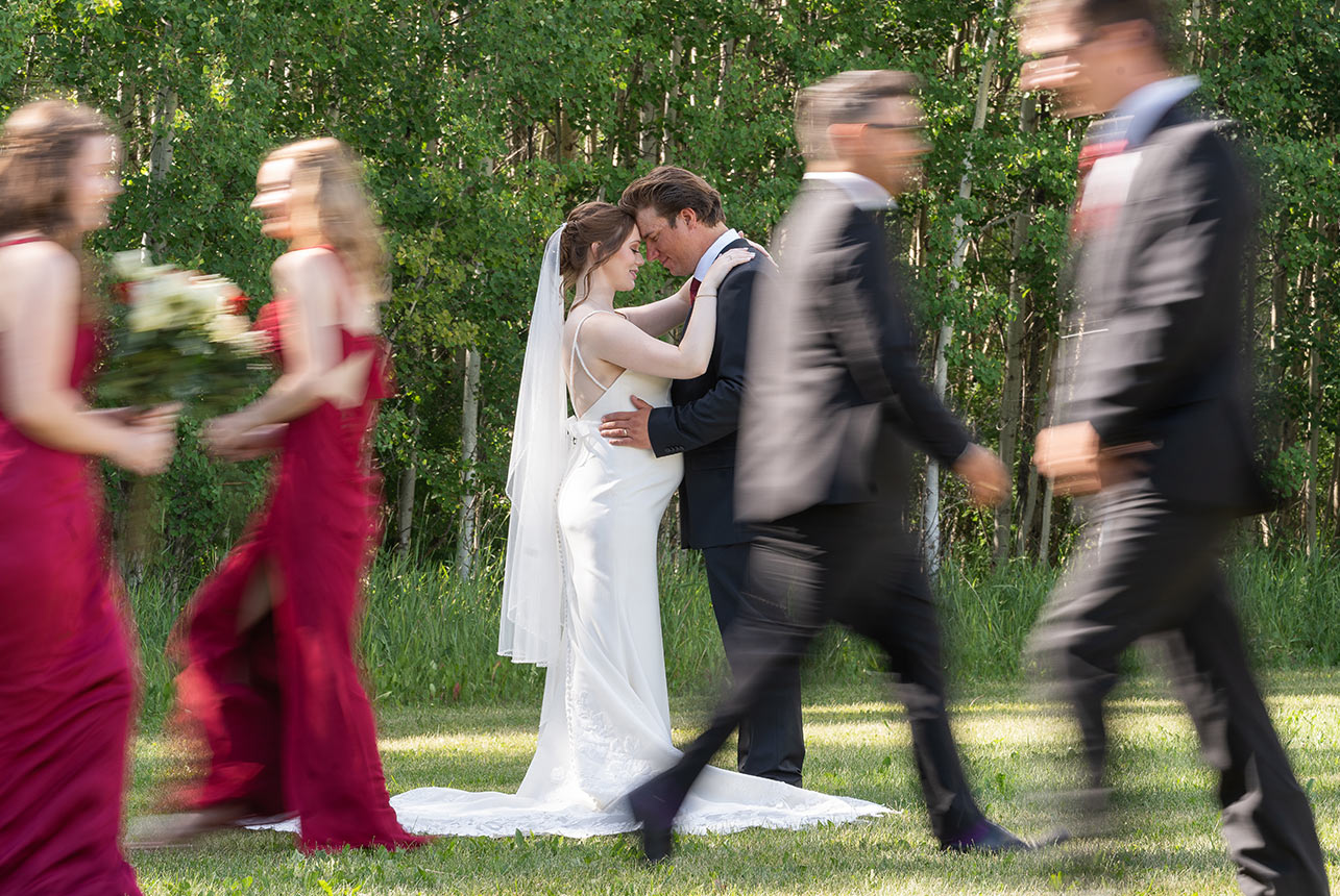 Bride and groom pause forehead to forehead, perfectly still, while bridesmaids and groomsmen blur past—an authentic, motion-filled wedding moment