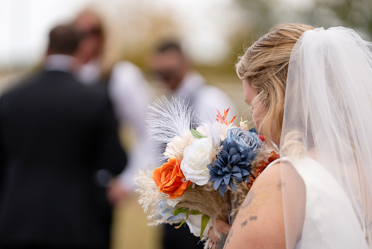 Bride’s backview as she smells her bouquet, with the groomsmen blurred in the background on a sunny autumn day