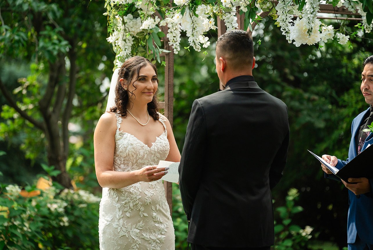 A bride with a joyful grin, basking in the sunshine, gazing lovingly at her groom, ready to read her notes during the wedding ceremony.