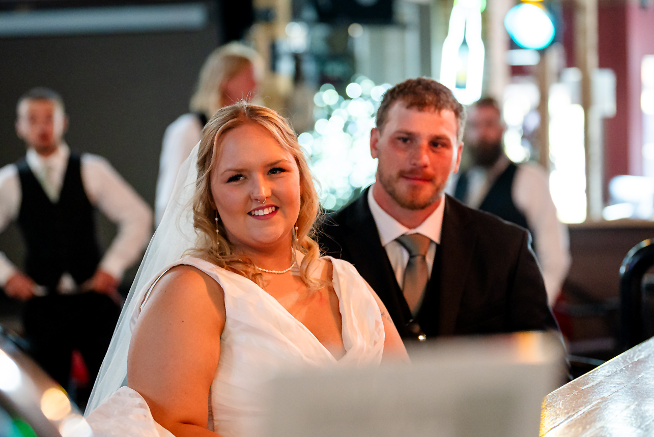 Bride and groom sitting together at the Kicks Saloon Bar in Redwater, with bokeh lighting and groomsmen in the background