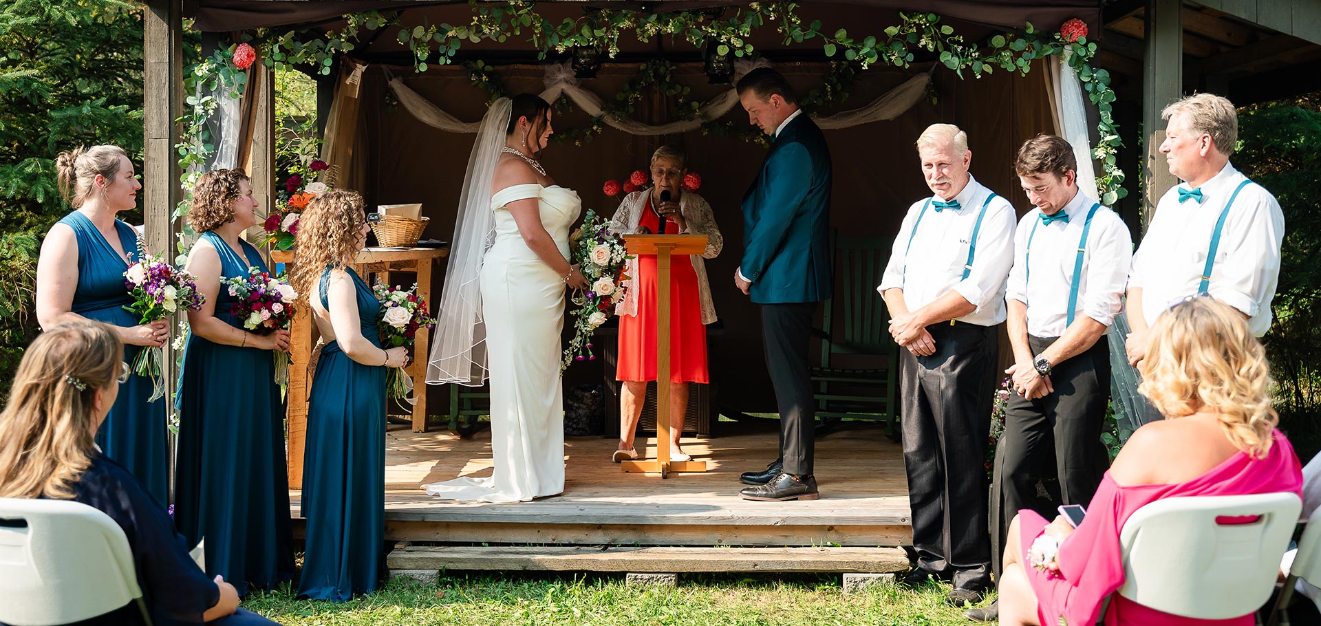 Bride and groom stand face to face on the ceremony stage, with bridesmaids and groomsmen aligned on the grass as the commissioner leads the vows