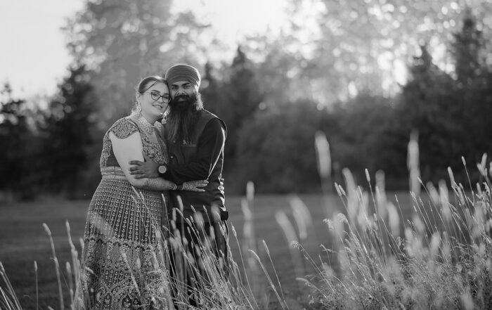 Black and white photo of the bride and groom standing in a wild grass field near Sherwood Park during golden hour, with trees in the backdrop.