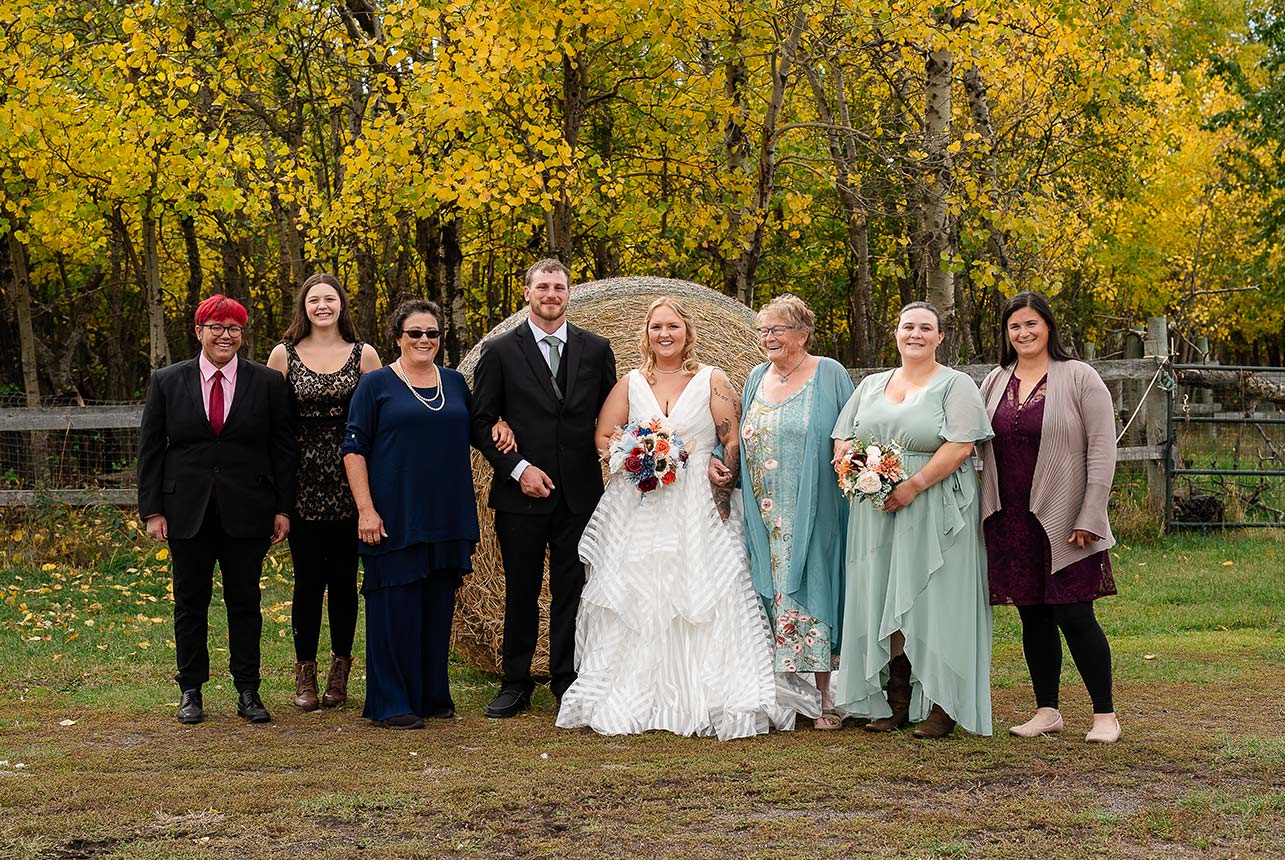 Bride and groom with guests posing for a group photo with haybales, a rustic fence, and autumn trees in Redwater, Alberta.