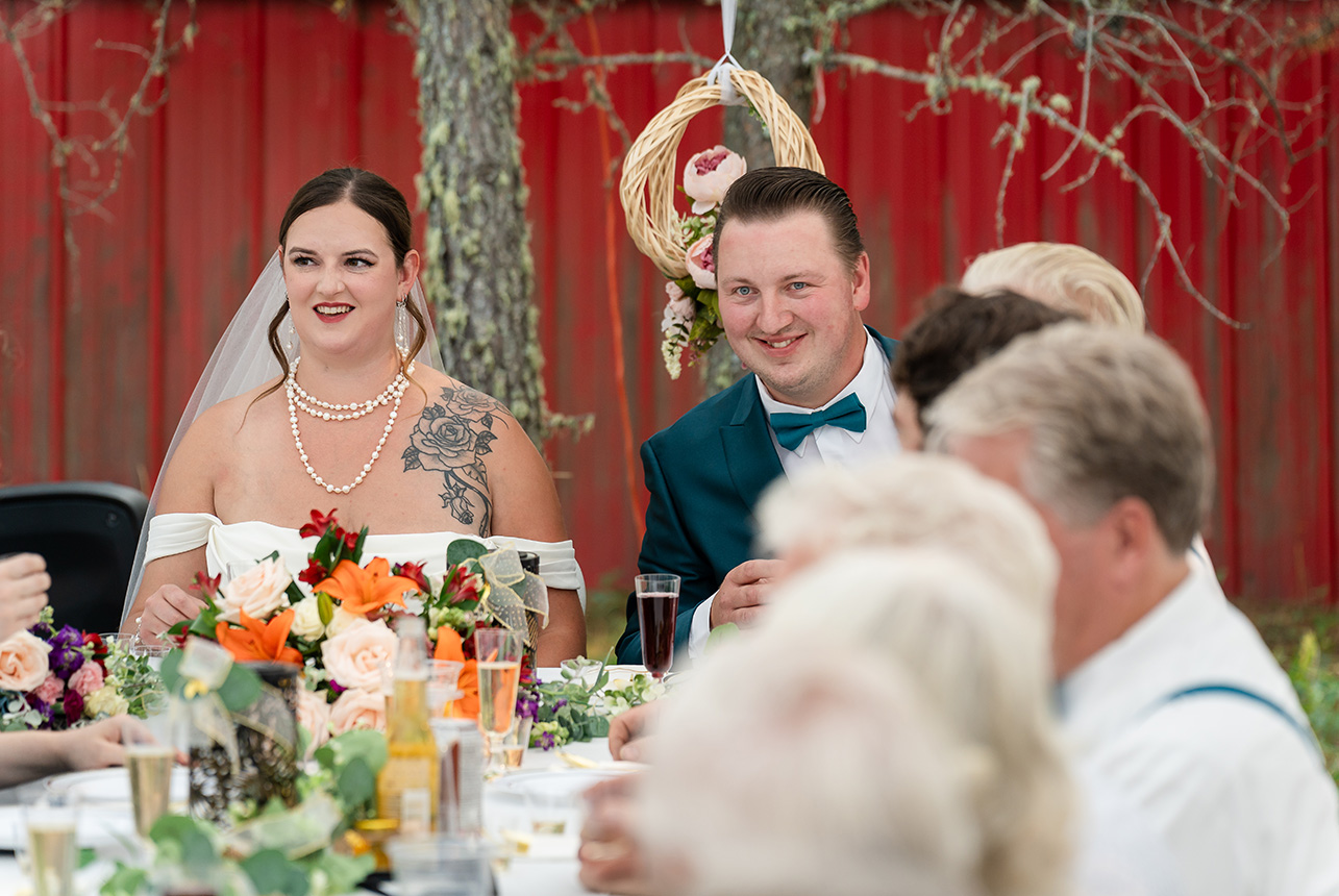 Bride and groom sit at the head of the reception table, sharing a moment together, with a red barn providing a perfect rustic backdrop