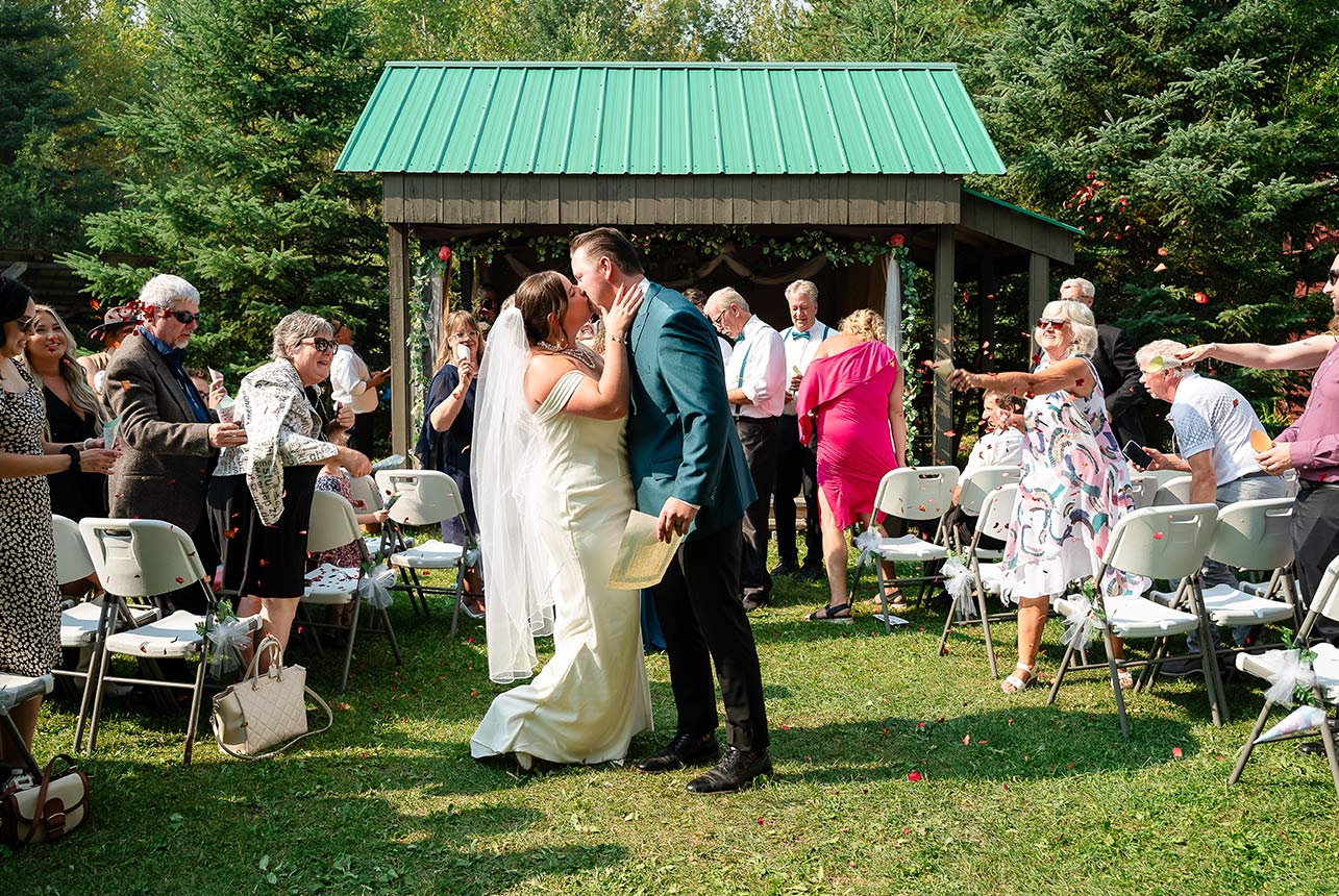 Bride and groom stop mid-aisle for a kiss as guests throw flower petals around them, celebrating their just-married moment