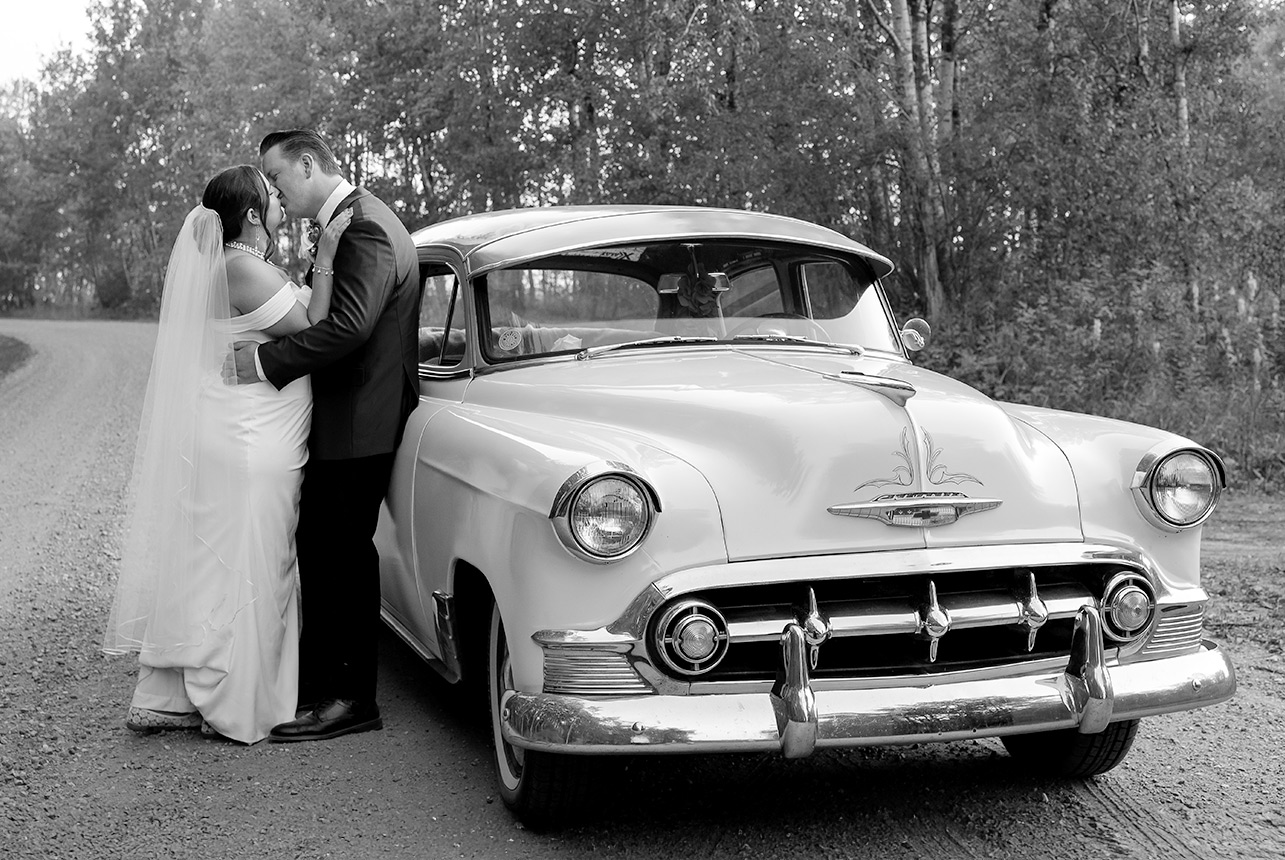 In a timeless black-and-white moment, the newlyweds share a romantic kiss on a curved gravel road, surrounded by the peaceful countryside