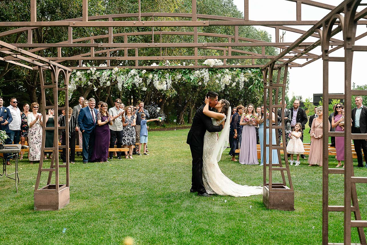 Bride and groom sharing a kiss behind the stage as guests applaud and children release bubbles during their wedding ceremony.