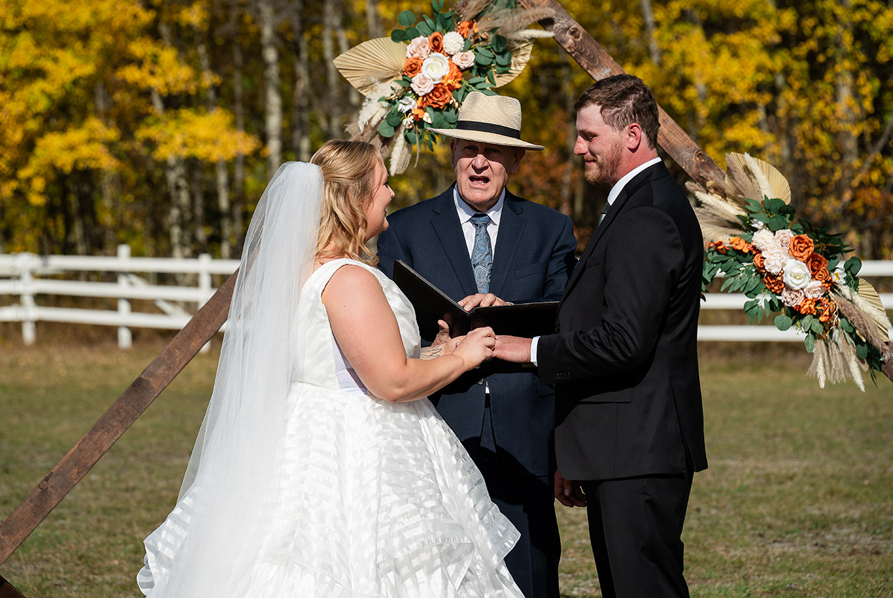 Bride slips the wedding ring onto the groom’s finger during vows at Finnegan Farms, surrounded by autumn trees and a white fence