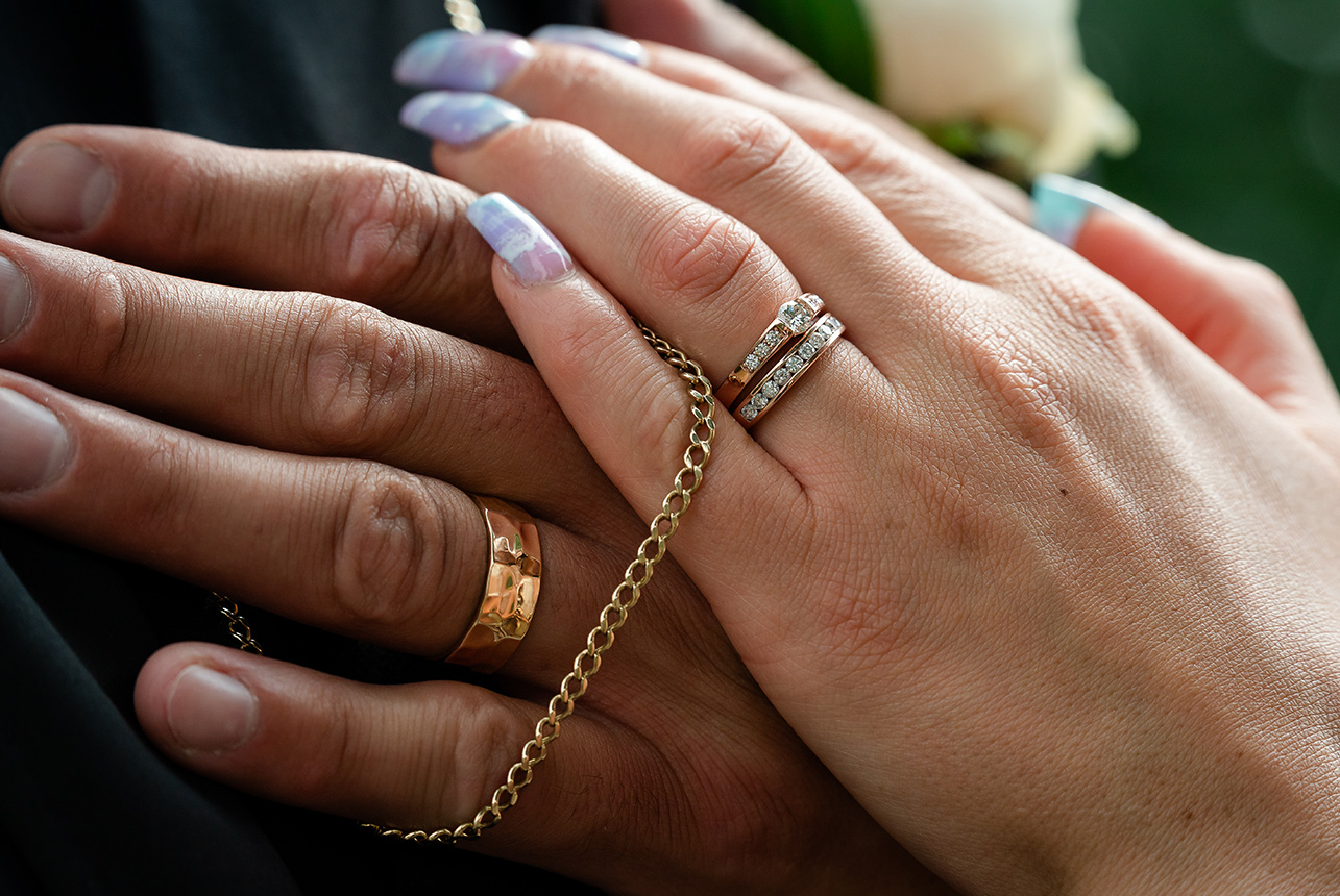 Close-up of the bride and groom's hands showcasing their wedding rings and the groom's gold chain woven between their fingers.
