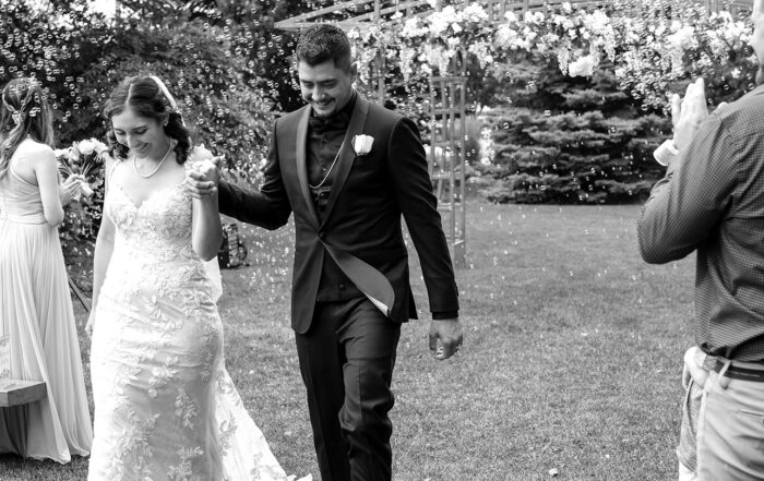 Black and white wedding photo of Juan and Hope walking hand in hand down the aisle at Greenland Botanic Gardens in Sherwood Park, near Edmonton, smiling with accomplishment and surrounded by small bubbles.