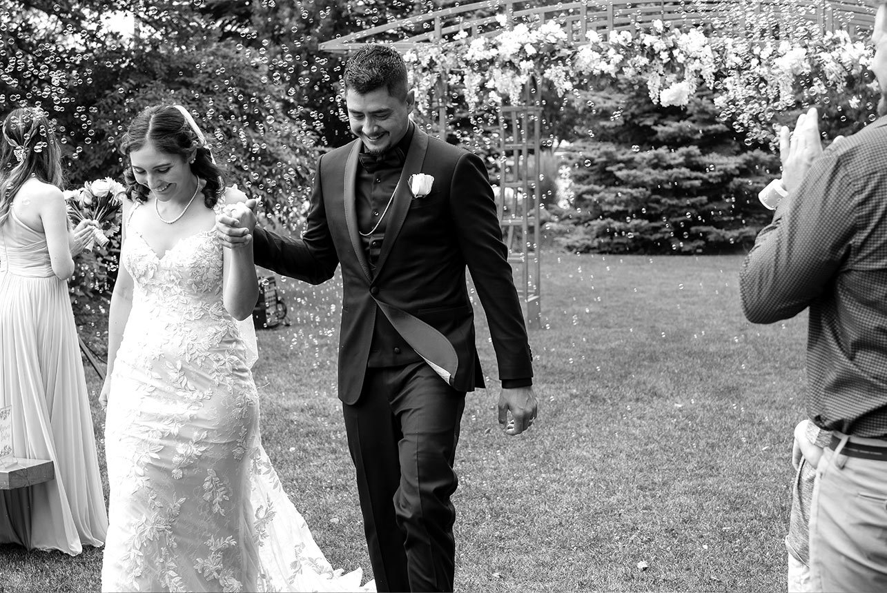 Black and white wedding photo of Juan and Hope walking hand in hand down the aisle at Greenland Botanic Gardens in Sherwood Park, near Edmonton, smiling with accomplishment and surrounded by small bubbles.