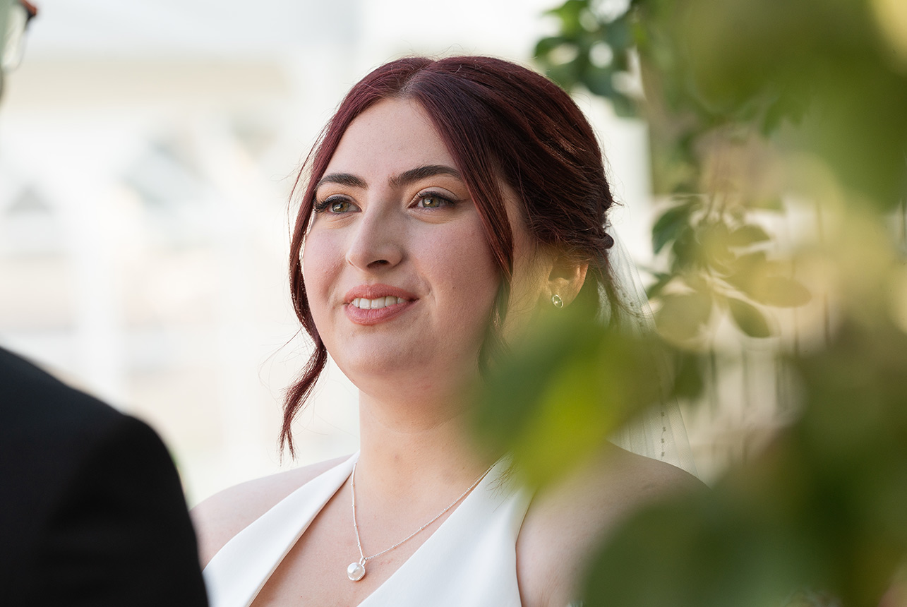 Bride listens to her partner during vows, soft garden light and gentle foreground leaves