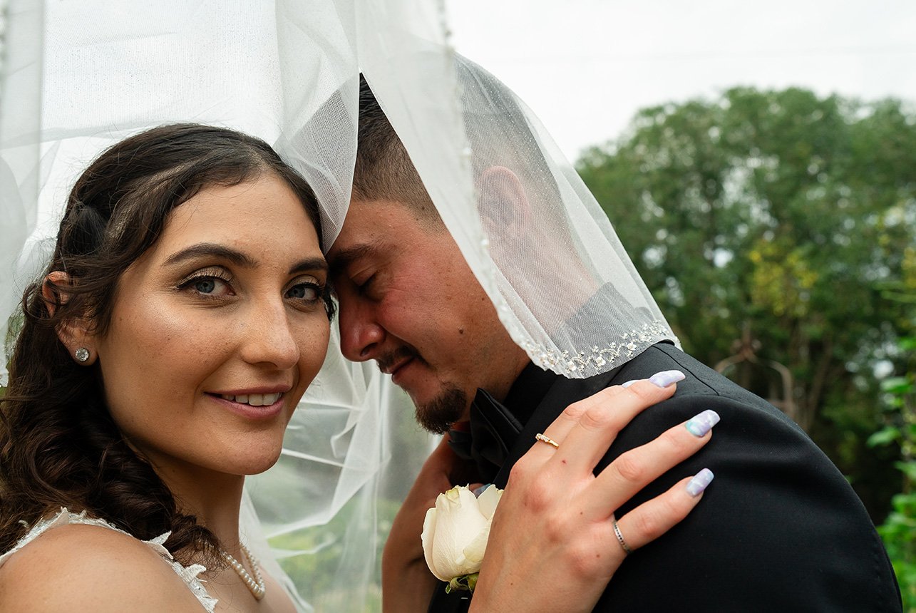 A bride looking towards the camera while her groom embraces her with his head bowed and eyes closed during their wedding ceremony.
