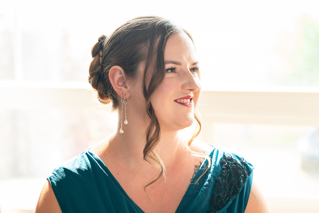 Bride stands near a window in soft natural light, her hair and makeup complete, preparing for the final touches before stepping into her gown