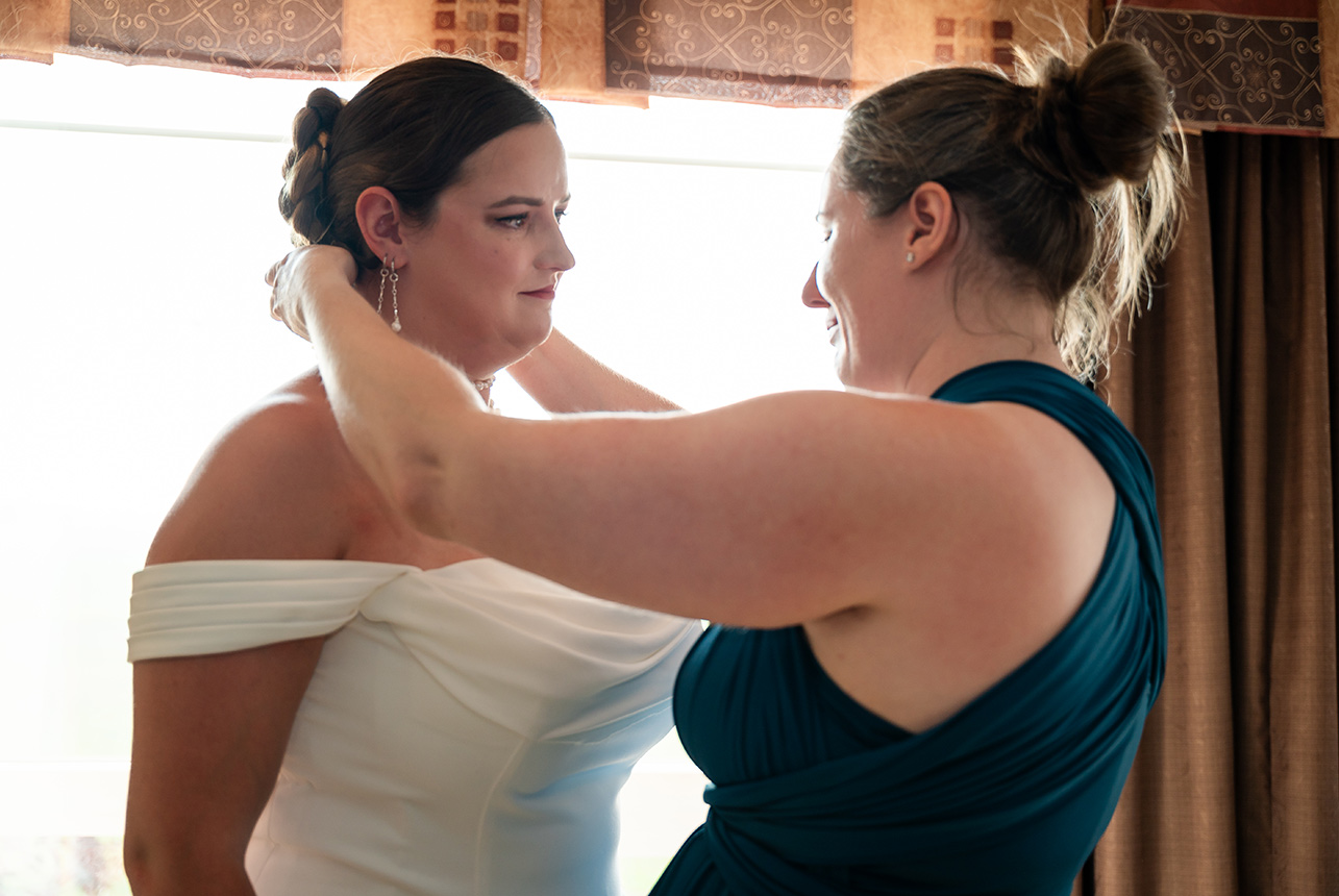 Bride’s sister fastens a pearl necklace around her neck, a heartfelt moment before she steps into her wedding gown