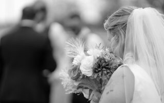 Bride pauses to smell her bouquet, surrounded by golden autumn leaves, with the groom and wedding party softly blurred in the background