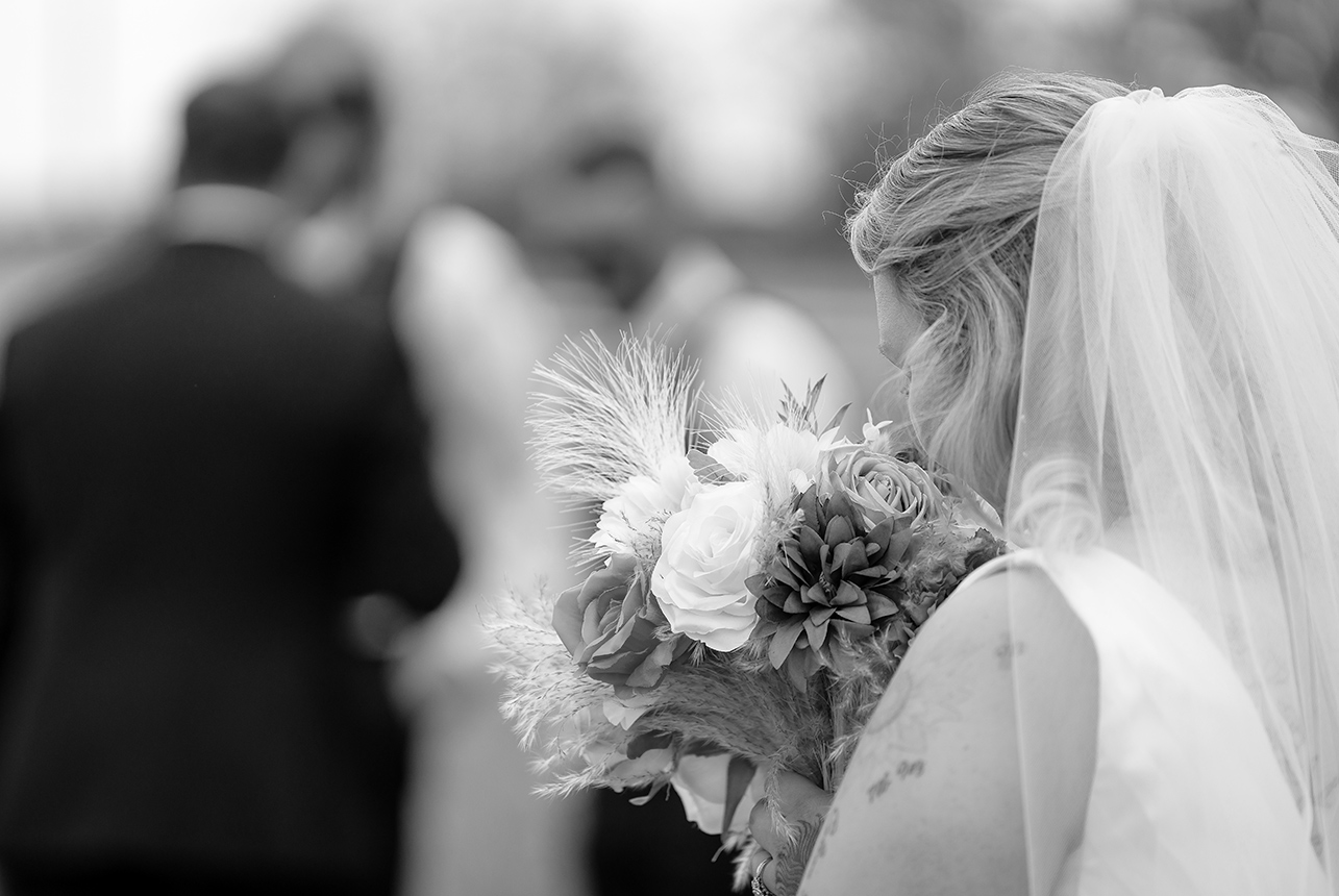 Bride pauses to smell her bouquet, surrounded by golden autumn leaves, with the groom and wedding party softly blurred in the background