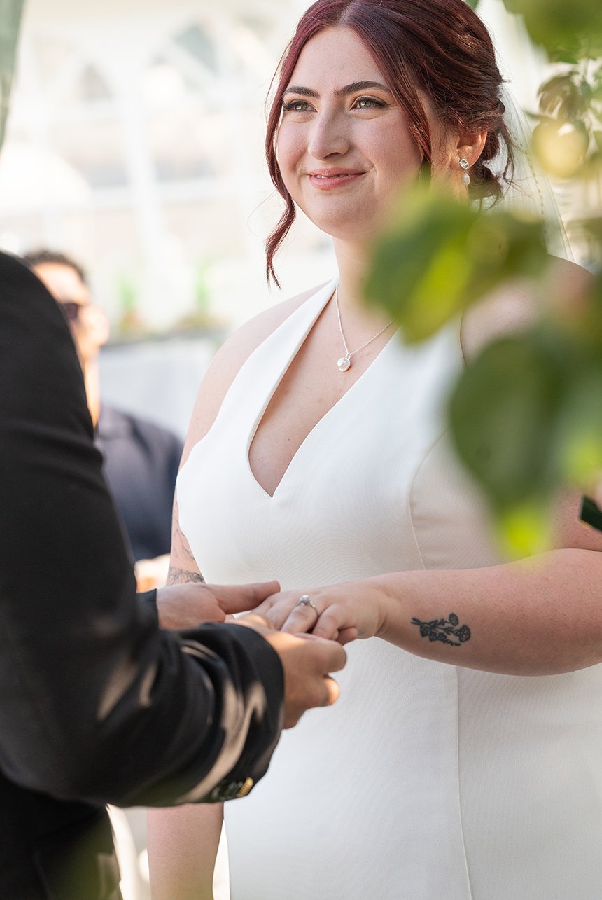 Bride smiling during the ring exchange, hands in focus with soft greenery in the foreground