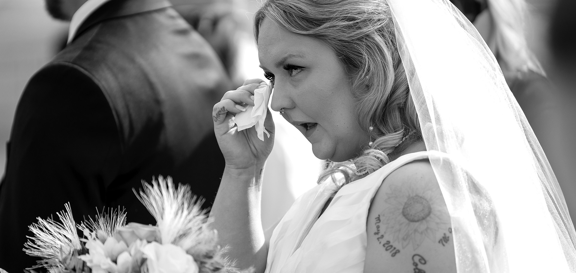 Bride wipes away a tear with a tissue, lost in an emotional moment amidst family and guests during her wedding, captured in timeless black and white.