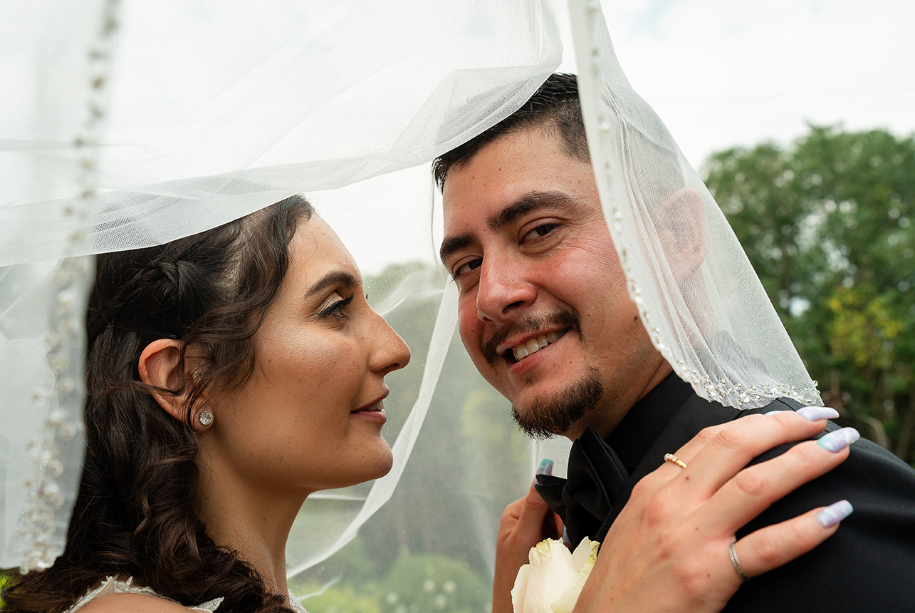 Bride under her veil with her hands on her groom's shoulders as he looks proudly towards the camera.