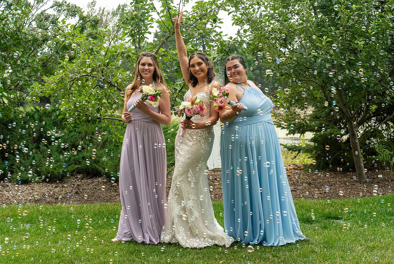 Bride standing between her two bridesmaids, making a V-for-victory pose with bubbles surrounding them, highlighting their dresses and holding bouquets.