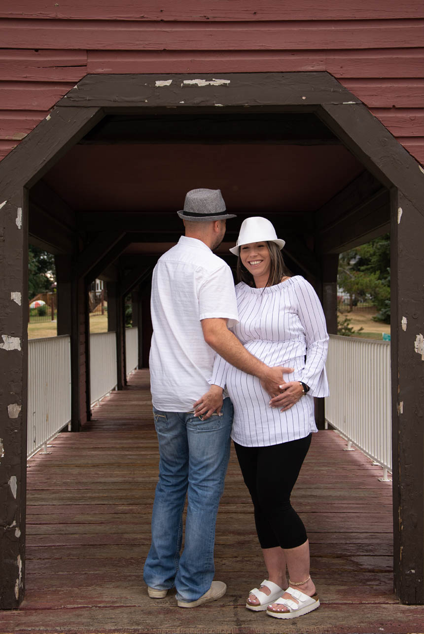 Expectant couple standing back-to-back on wooden bridge in Sherwood Park, showcasing their love and anticipation for their growing family.