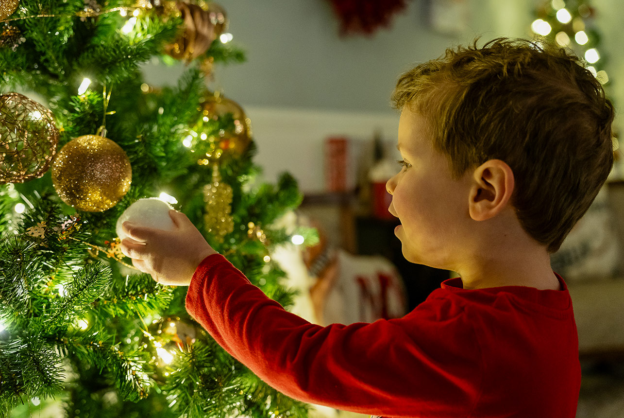 Young boy in red pajamas placing ornament on a glowing Christmas tree indoors