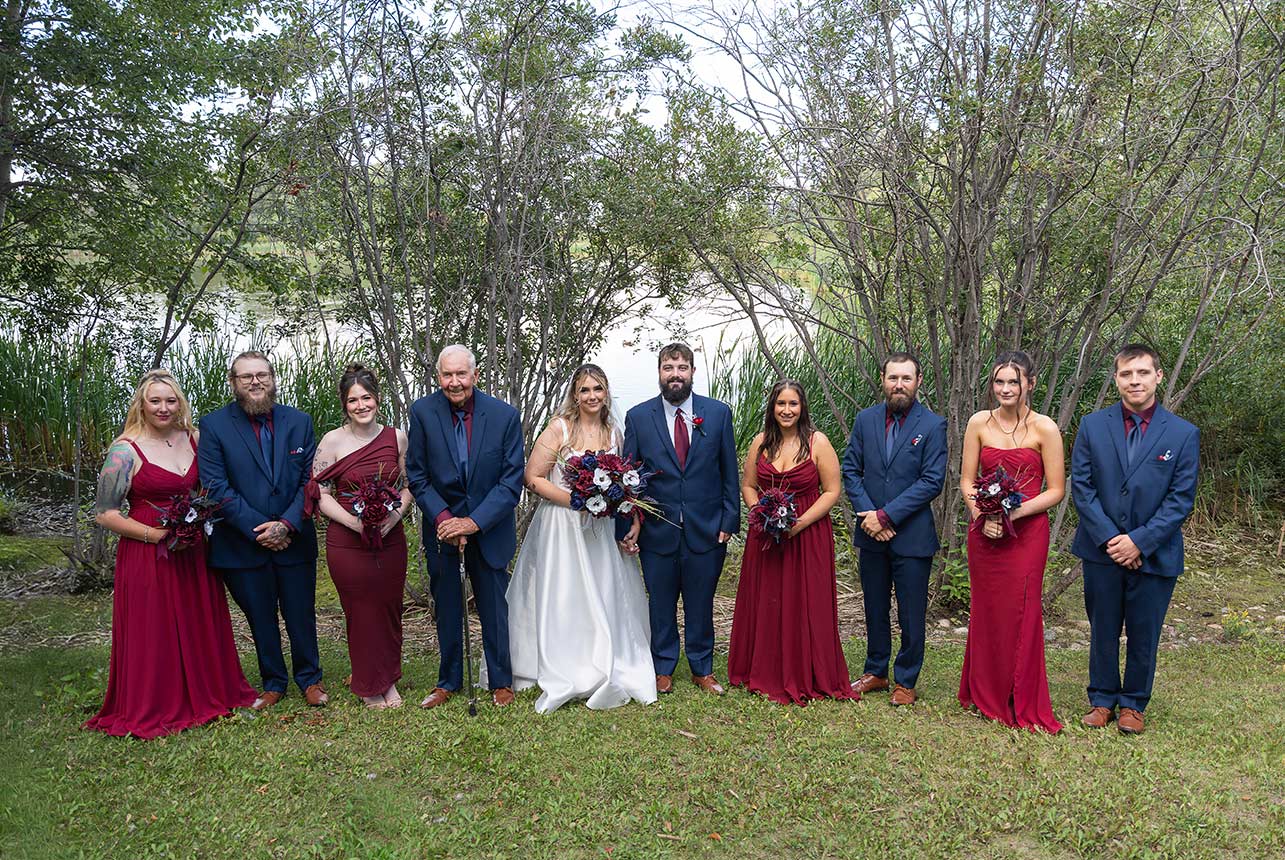 Wedding party portrait outdoors by the water with the couple centered, bridesmaids in burgundy and groomsmen in navy, framed by trees