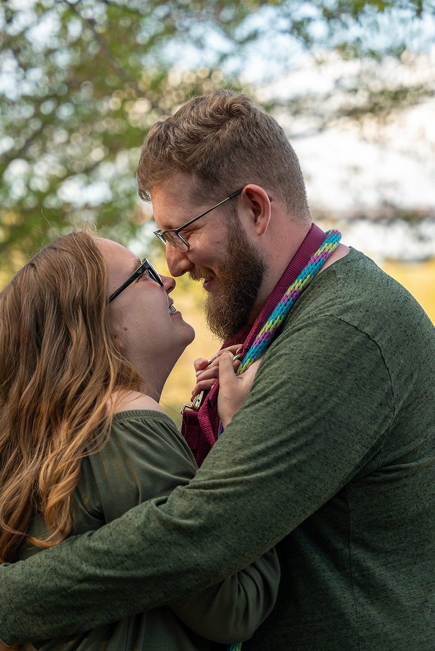 Engaged couple smiling closely and touching noses, surrounded by trees and soft evening light
