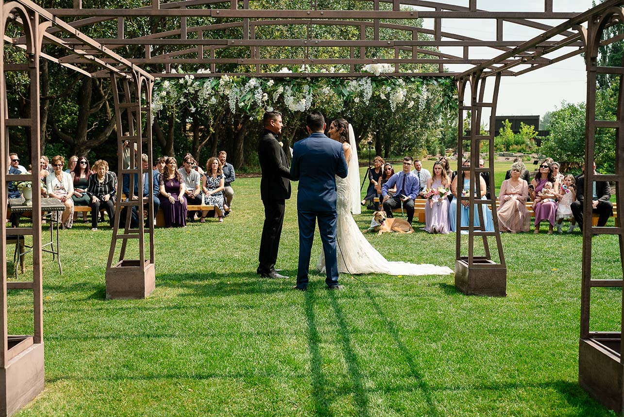 A back view of the commissioner reading vows to the bride and groom during their wedding ceremony at Greenland Botanic Gardens, with family and guests in the background.