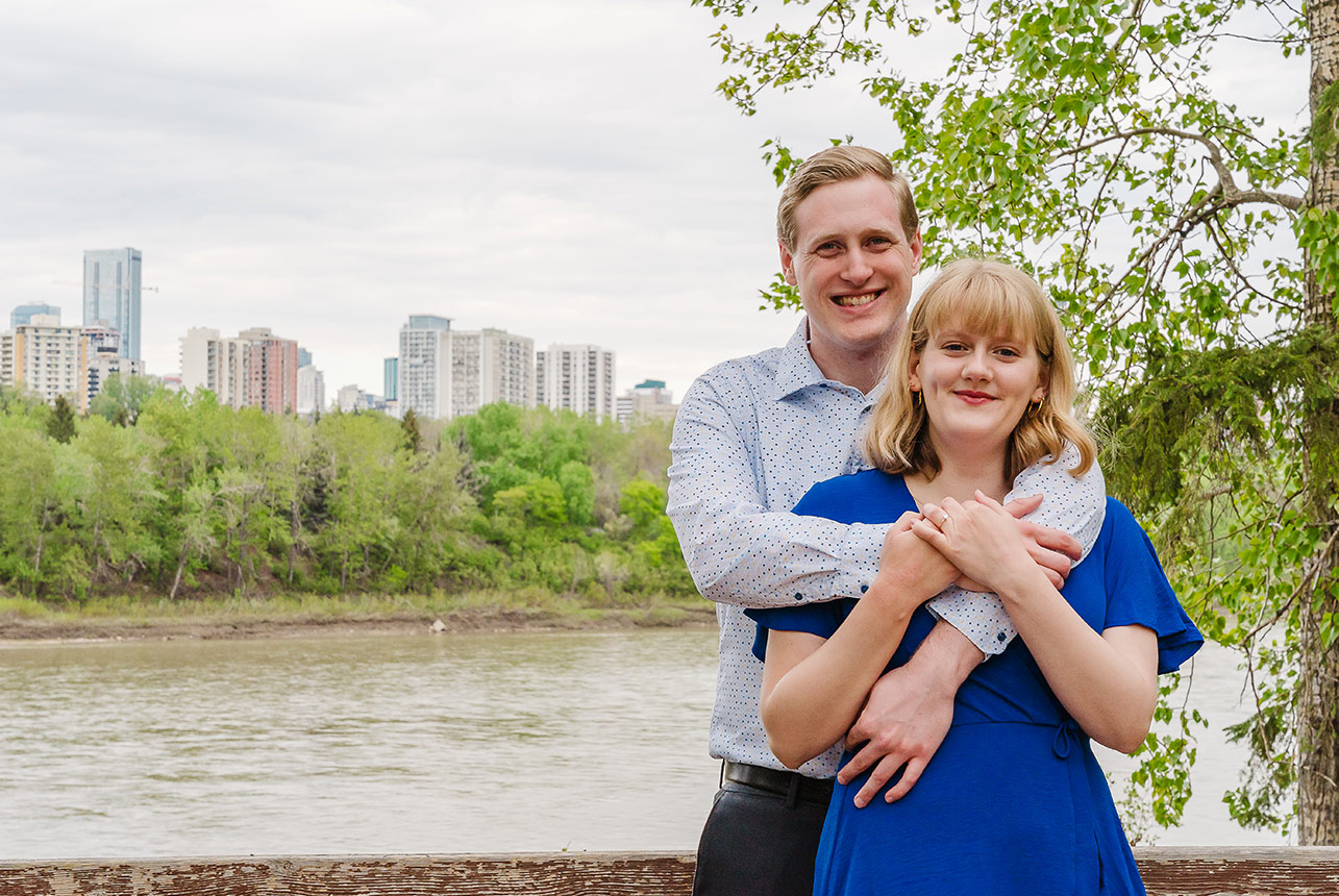 Couple with woman in a blue dress, standing by a river with Edmonton's skyline in the backdrop.