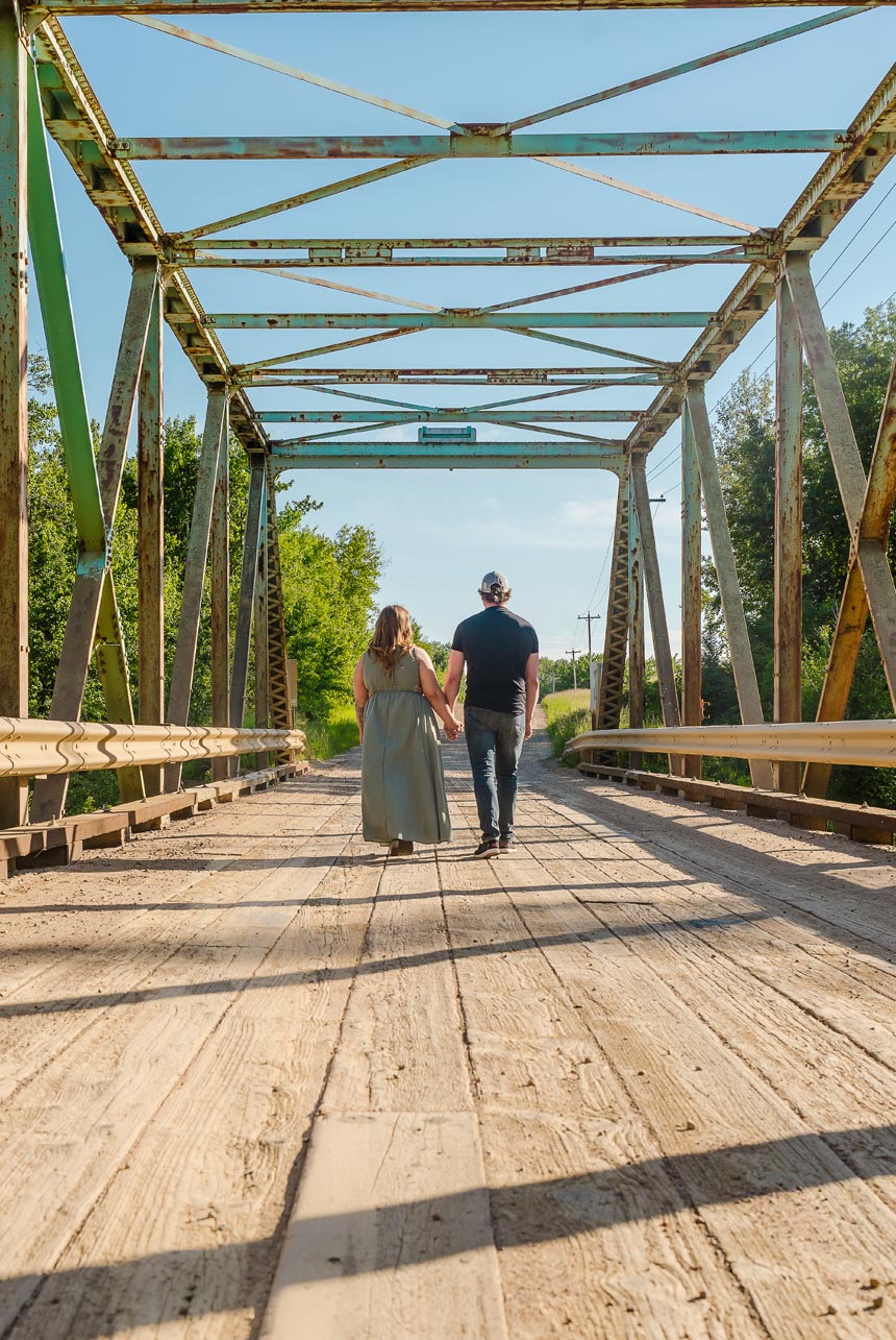 Couple holding hands walking across a rustic steel bridge on a sunny evening in Redwater, Alberta