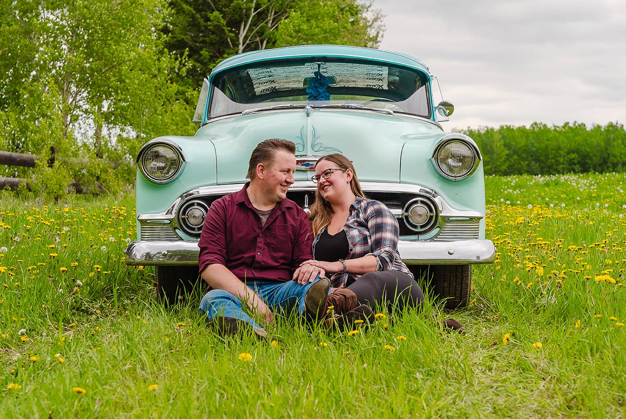 Couple sitting in tall grass by antique car in Edmonton field with dandelions, country fence, and forest in the background.