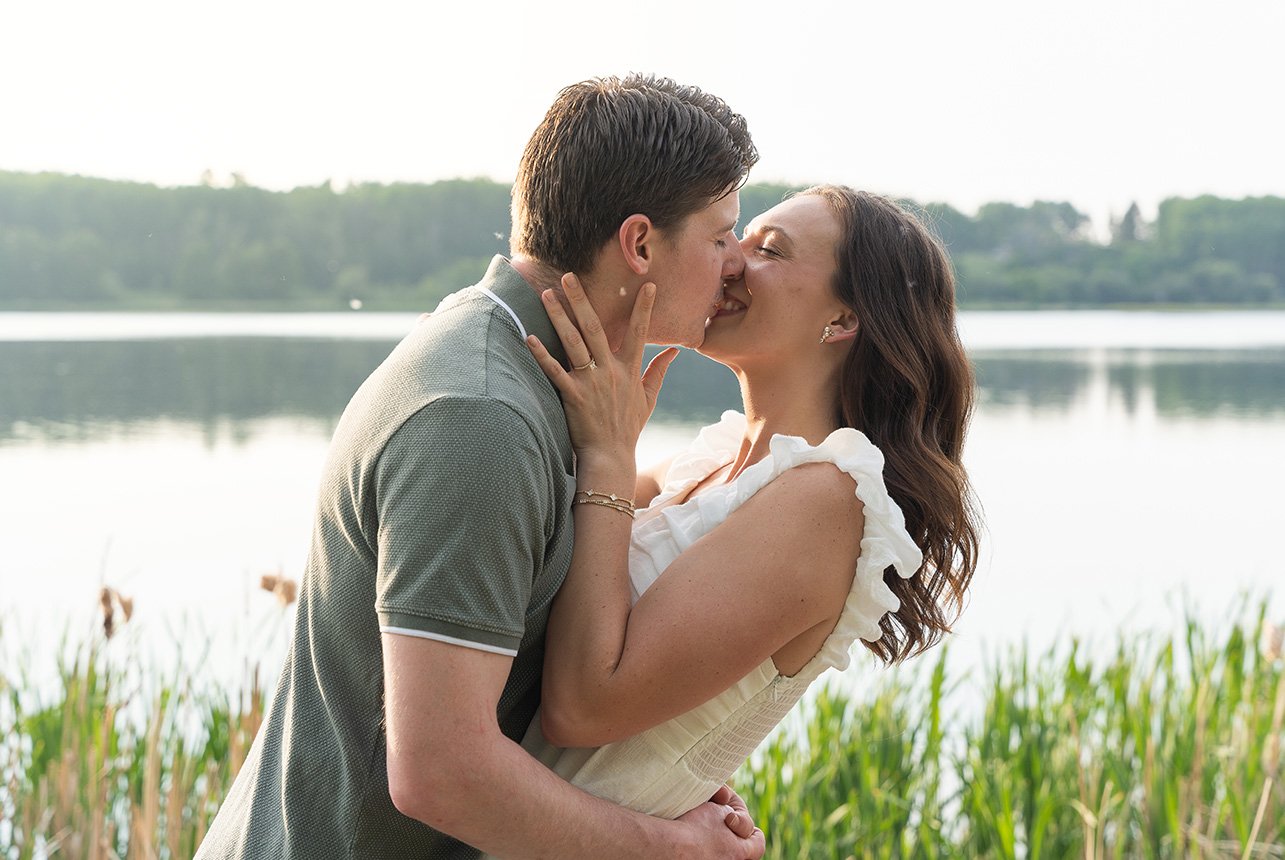 couple kissing by private alberta lake Couple shares a sweet kiss in front of a still lake, surrounded by tall grasses and soft summer light