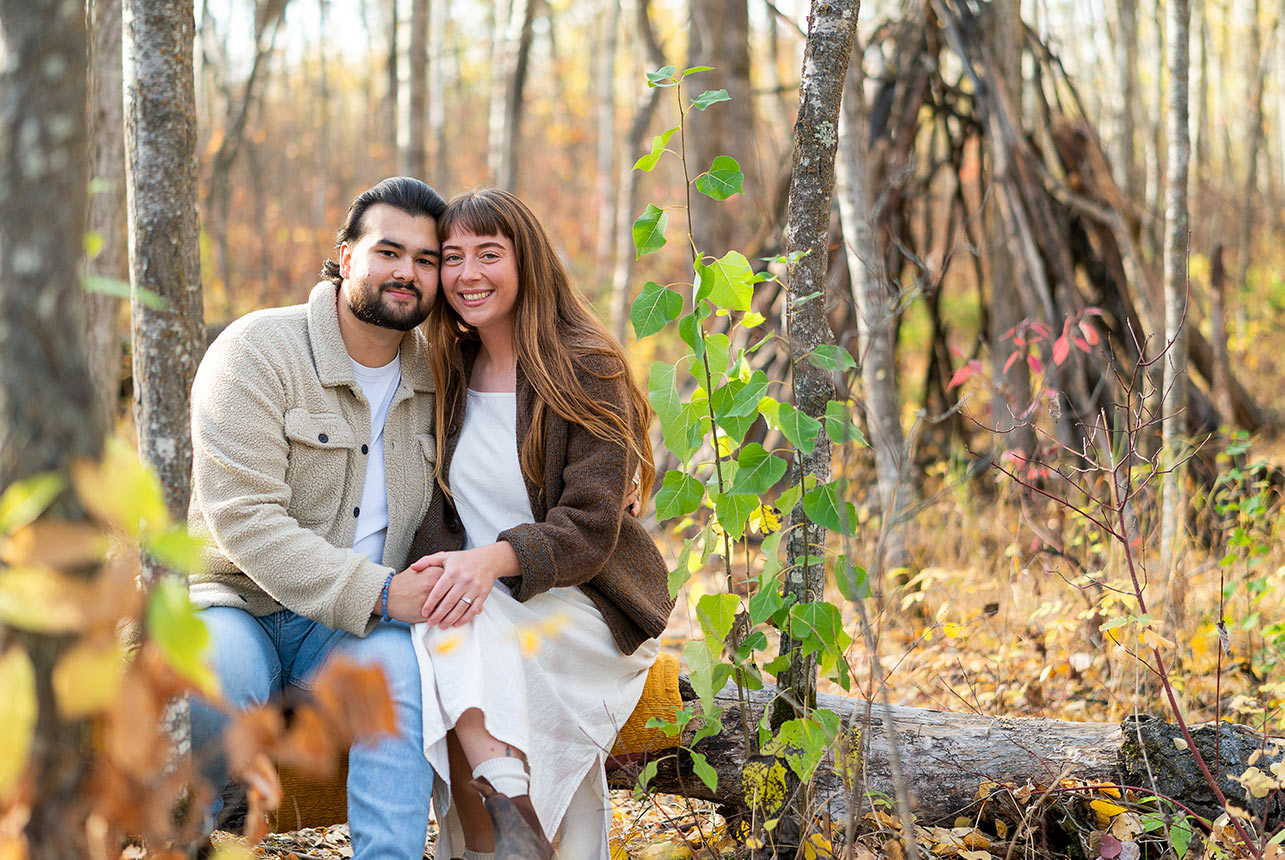 Engaged couple sit together on a fallen log at the Sherwood Park Natural Area, smiling in soft autumn light