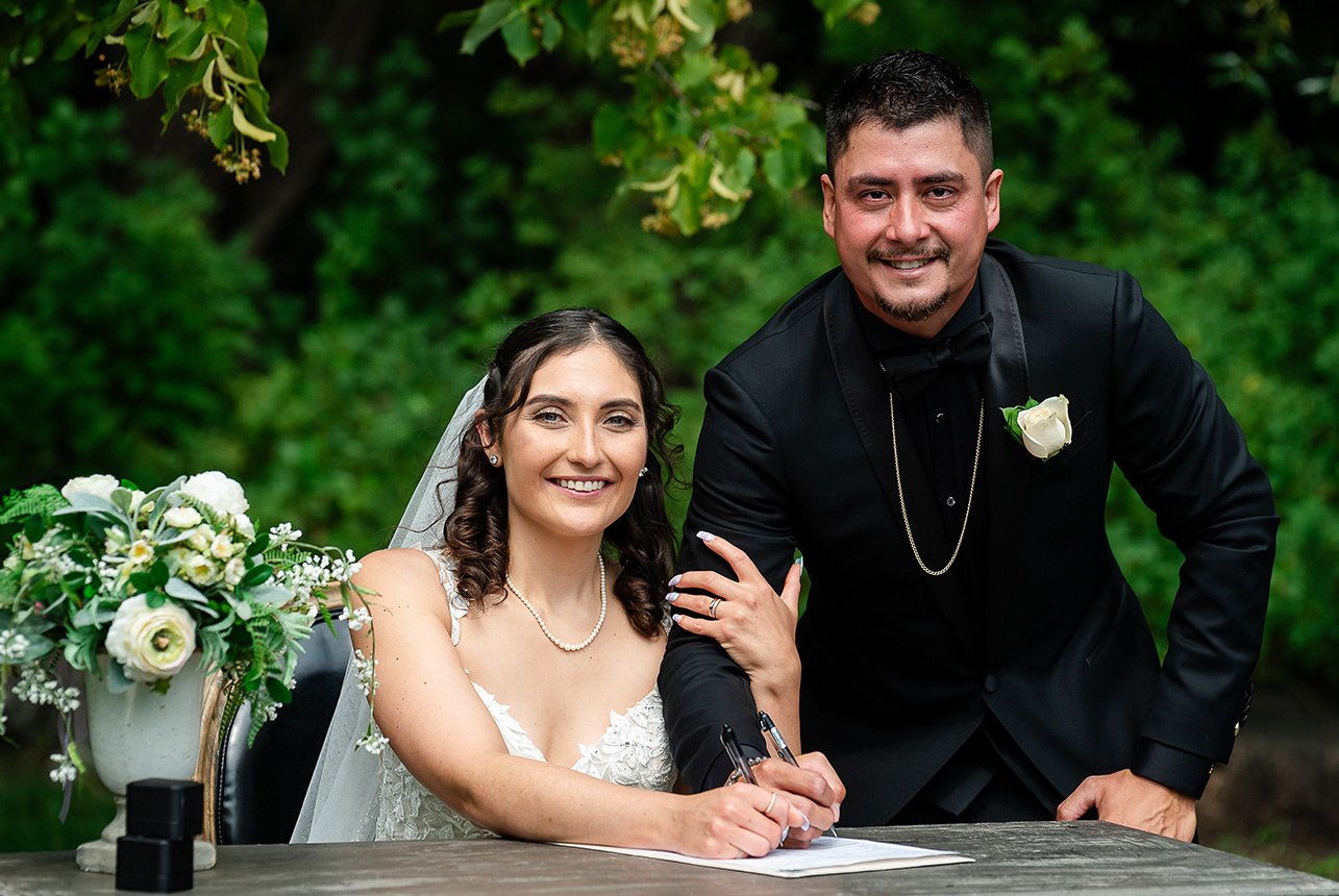 Bride seated displaying her wedding ring while the groom signs the marriage contract at an outdoor wedding in Edmonton.