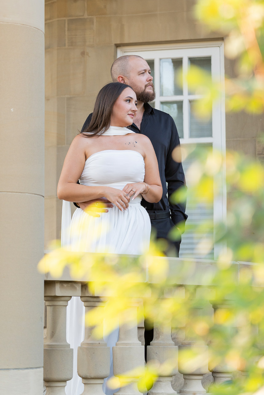 Engaged couple on the balcony at Government House in Edmonton, framed by soft foliage as they look out together