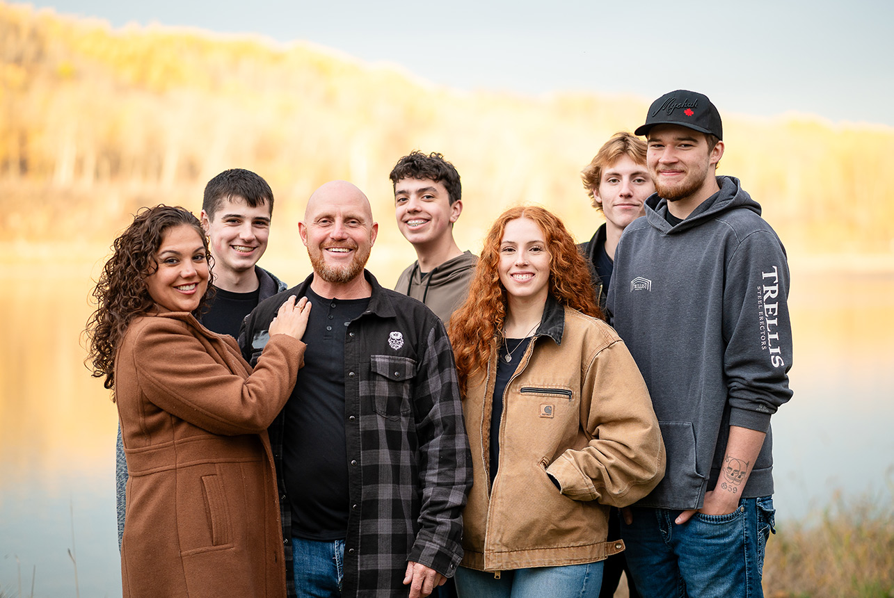 fall lake family portrait five teens Large blended family with seven teenagers poses by a lake at sunset during a fall portrait session in Devon Alberta