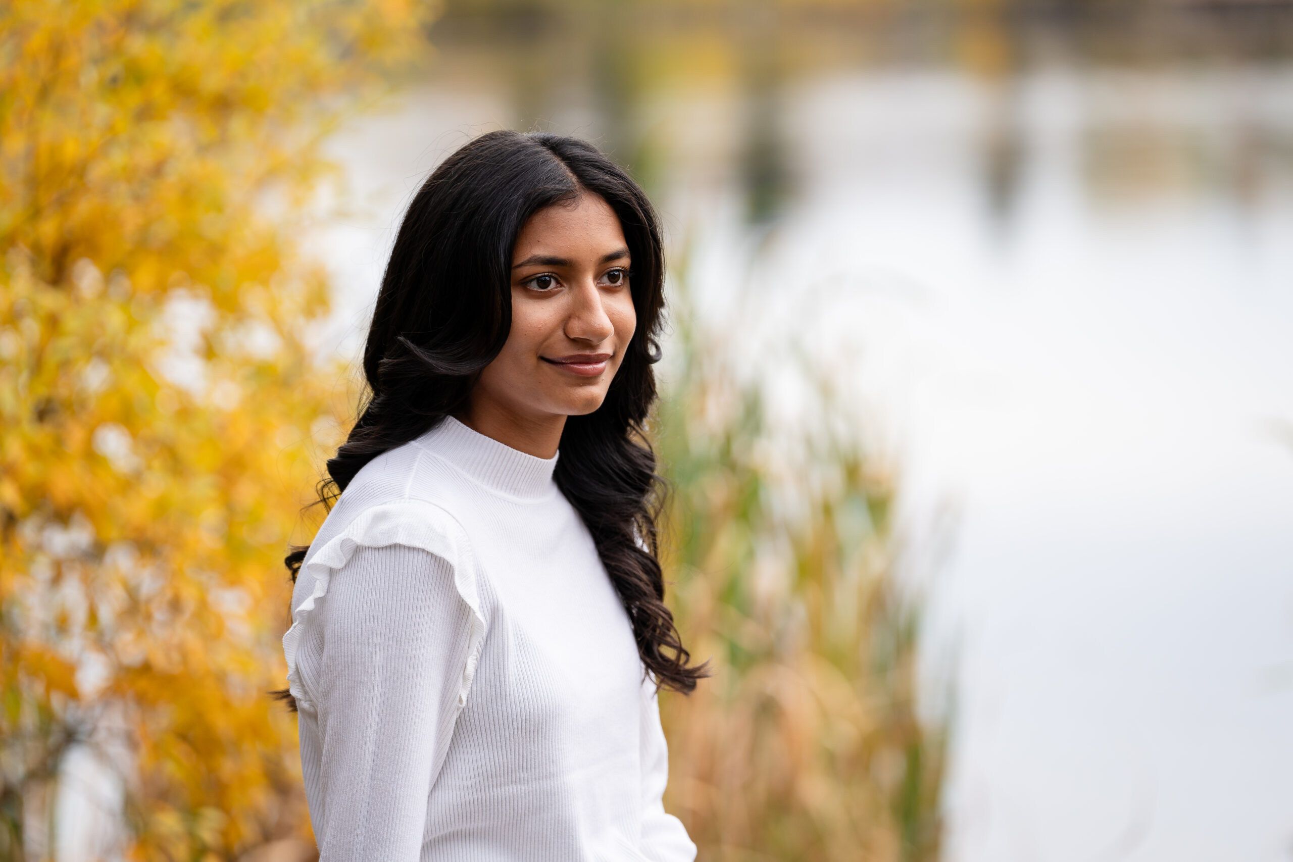 Teen girl in white sweater smiling softly beside golden foliage and a calm lake in Edmonton