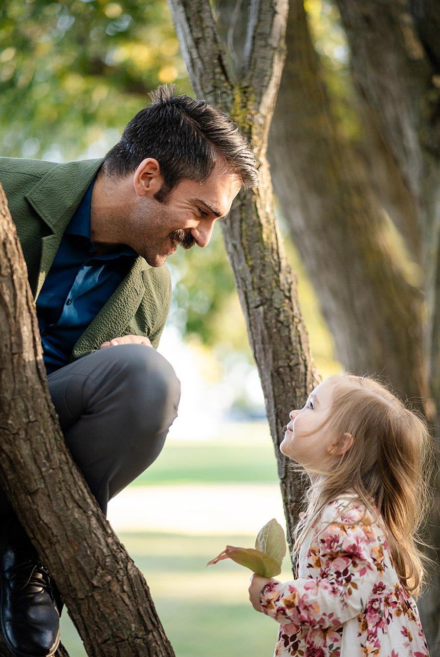Smiling father crouches in a tree talking to his young daughter holding leaves at an autumn park photo session in Edmonton