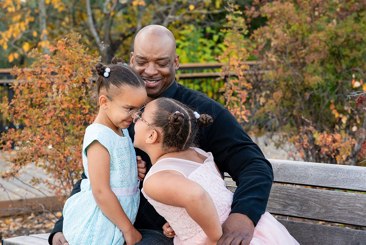 Father sitting on bench smiling as his two young daughters in dresses share a playful moment