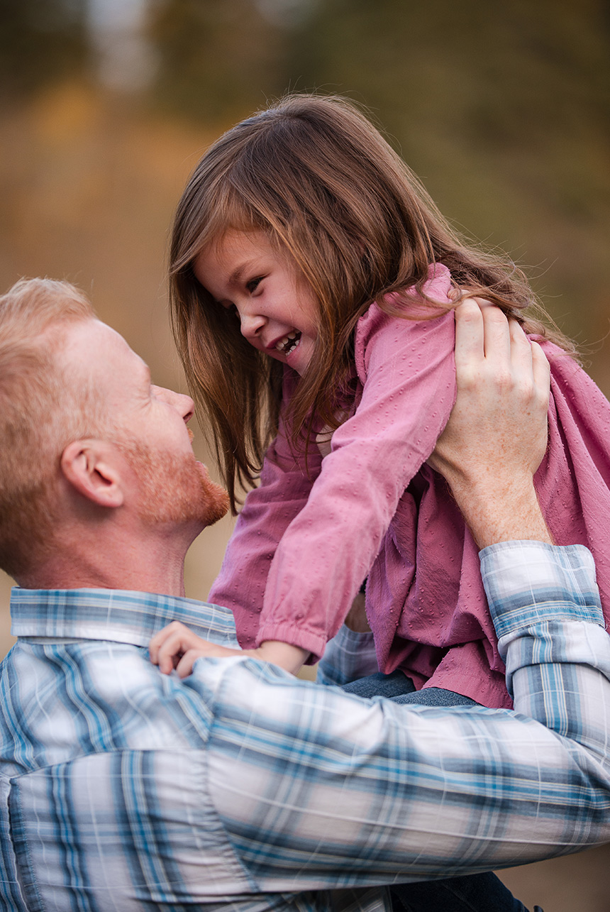 Father lifting his toddler daughter high in his arms as she gazes at him, surrounded by autumn colors in an Edmonton park.