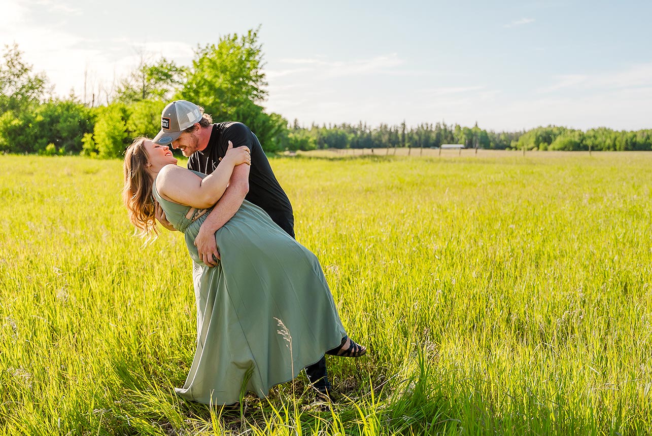 Groom-to-be dips his fiancée for a kiss in a glowing summer field with sunshine spilling over them