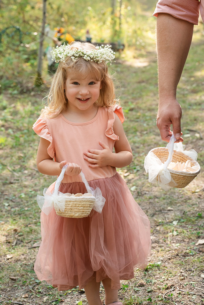 Flower girl in a peach dress and white floral crown walks up an enchanted forest path, carrying a wicker basket toward the wedding aisle