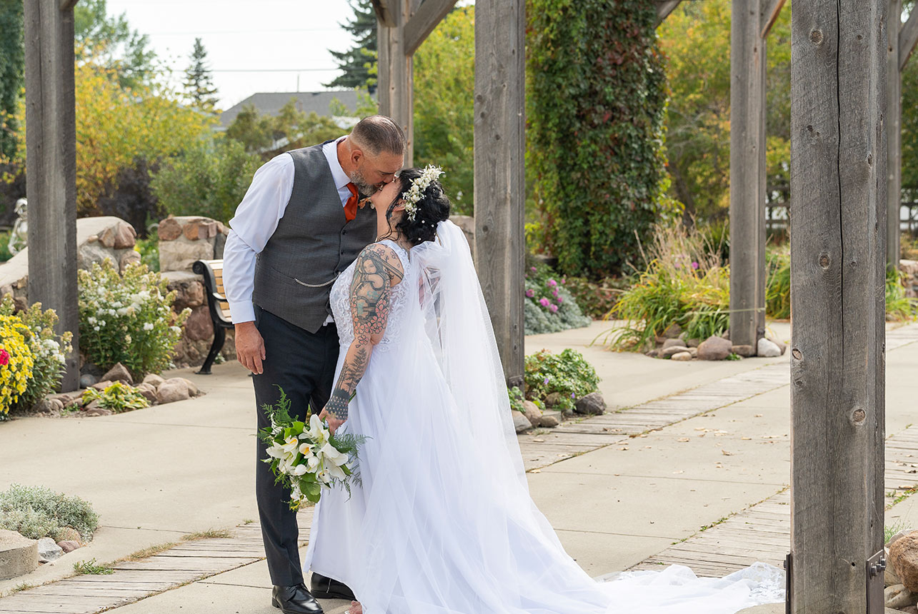 Newlyweds share a kiss beneath a wooden pergola, bouquet of lilies and a flowing veil