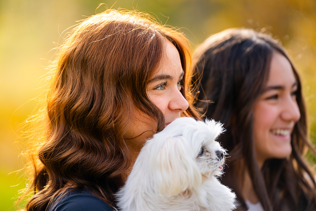 Teen sisters laugh while one cuddles a fluffy white Maltese during a golden‑hour family session in Edmonton