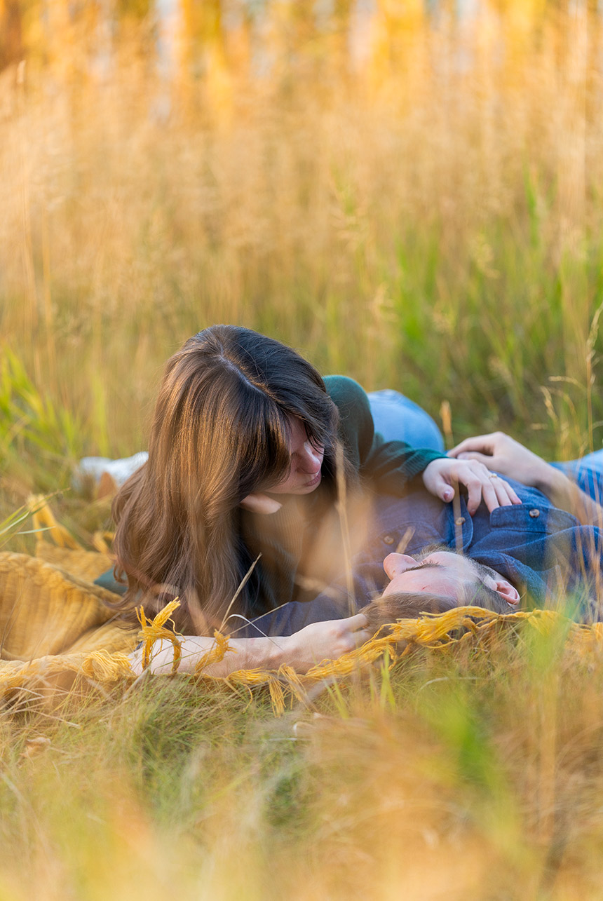Couple reclining on a mustard blanket in tall golden grass, sharing a quiet gaze while holding hands at sunset